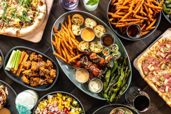 Selection of menu items featuring The Sampler sharing platter, Pan-Roasted Shisito Peppers, The Spicy Hawiian pizza, and Hot Honey Sweet Potato Fries.