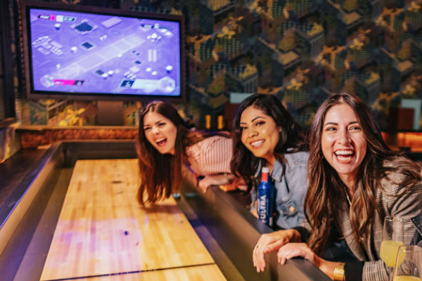 Three friends leaning over an Electric Shuffleboard table in the Miami Room during brunch.